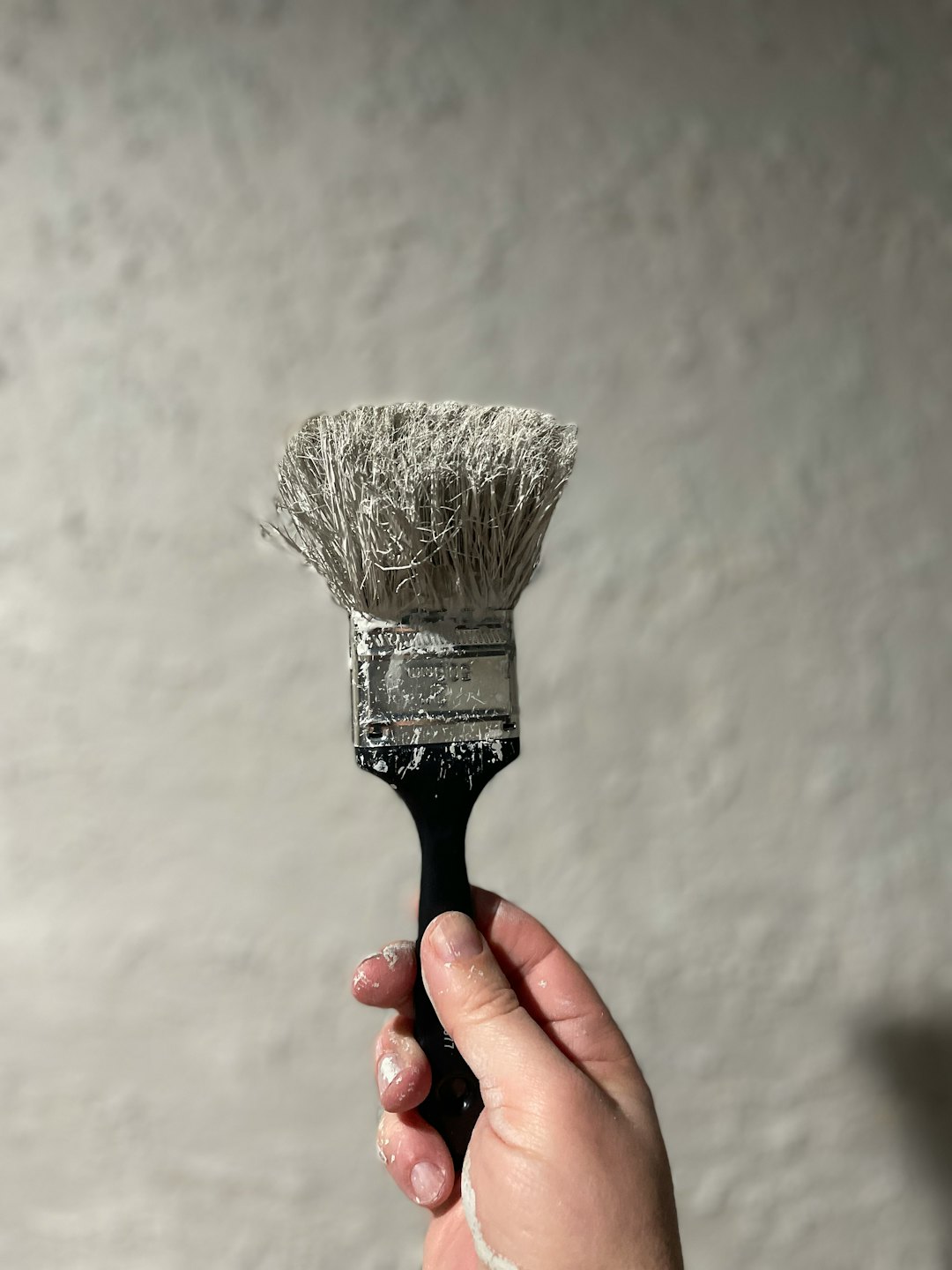 Hand mit weißer Farbe hält einen bemalten Pinsel vor eine Wand.